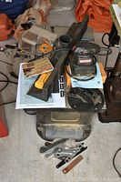Top view of the router table components including trapezoid router plate, dust port, mounting hardware, and hand sander placed on top, along with manuals and packaging.