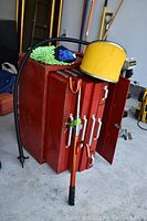 Red metal tool chest with open side door, white handles, yellow foot pump on top, green cleaning mitt, blue cloth, orange garden tool leaning against it, and black hose connected to foot pump.