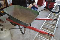 Full side view of the wheelbarrow showing green metal tub, red wooden handles, black metal legs, placed on a garage floor with various items around.