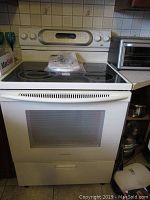 Front view of white KitchenAid electric stove showing smooth ceramic cooktop, oven door with viewing window, control panel with dials and display, and manual placed on top.