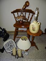 Wooden barstool with lamps and other items on and around it, small wooden table beside it, electric fan and candles on floor.