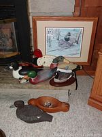 Photo showing multiple wooden duck decoys arranged on floor and hearth, with framed duck print leaning in background.
