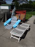 Photo showing two older web lawn chairs, floating pool lounger with blue resin parts, white adjustable plastic lounger, and wrapped blue patio umbrella on concrete patio.