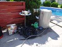 Photo showing beige rain barrel with hose, green watering can, black hose coiled and some garden decor items including metal butterfly decoration and birdbath base.
