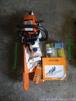 Top-down overview showing chainsaw with bar cover, manual, spare chain and oil bottles