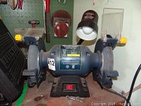 Front view of the blue Mastercraft 8 inch bench grinder with dual grinding wheels, mounted on a black base. The grinding wheels have protective guards, with a clear shield on the right side. A small wrench is placed near the front base.