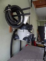 Side view of the small shop vacuum mounted on a wooden bracket with coiled black hose attached and extra bag visible.