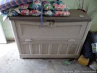Photo of beige resin deck box in closed position with floral cushions on top, showing front and top surfaces.