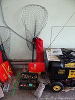 Wide view showing large fishing net with red carrying bag, tackle box with various lures, and portion of lawn chair and other items beside a generator.