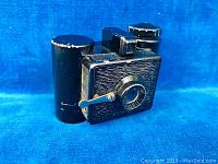 Front angle view showing black textured die-cast camera with crank, lens and cylindrical metal case attached on the left side.