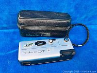 One Minolta 16 QT camera in silver with black leather carrying case and wrist strap on blue background.