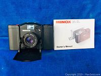 Front view of black Minox 35 EL camera with lens cover flap open, showing the Color-Minox 1:2.8 40mm lens and attached owner's manual booklet.