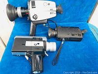 Three vintage movie cameras arranged on a blue cloth background showing front and side views.