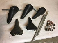 Photo showing five bicycle seats, two metal air pumps, and small collection of bicycle parts including clamps and brake pads on a white surface