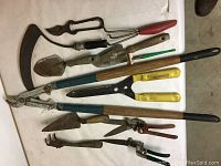 Photo of various garden tools laid on a white cloth, showing two loppers, hedge trimmer, grass clippers, weed puller, spades, sickle and snipper.