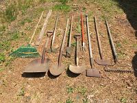Photo showing all garden tools laid out on grass and dirt, including green plastic grass rake, metal rake, shovels, hoes, pitch fork, scraper, and level.