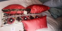 Four throw pillows on bed including two solid red pillows, one long decorative pillow with geometric red, white, brown, and black pattern, and one red pillow with ribbon ties.