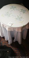 Round table covered by white fabric embroidered with blue and green floral patterns, topped by clear glass.