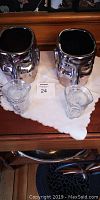 Two silver-tone face-shaped flower planters and two clear glass face cups placed on a white cloth on wooden surface.