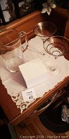 Set of two spiral stemmed glasses, clear glass pitcher, and a white recipe box on a tabletop with a decorative cloth.