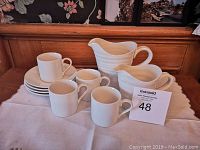 Photo of two creamers, four espresso cups, and four saucers on cloth-covered surface with wood background.