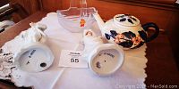 Old ceramic teapot with lid beside lead crystal candy dish and two porcelain figurines on a lace cloth