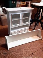Front view of white wooden hanging cabinet with glass doors and bottom drawers, alongside white hanging shelf placed on floor.