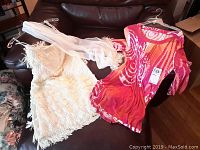 Overall lot showing both cream fringe tunic and red-pink flapper style dress with scarf laid on a couch.