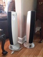 Two Sharper Image Ionic Breeze Ionic Breeze air purifiers standing side by side on wooden floor, against a wall, showing front and side views with cords wrapped at base.