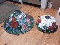 Two stained glass lamp shades placed on wooden floor. Larger shade features multicolor floral pattern, smaller has white background with red flowers at the border.