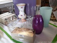 Photo of seven loose vases including floral ceramic, purple glass, clear glass varieties, and blue-green vase on a white surface near a window.