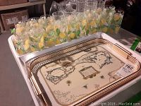 Plastic lemon-patterned cups arranged on the large trays. 10 tall and 10 shorter lemon cups are visible plus clear cups behind. The smaller Bermuda map tray is visible under the gold-trimmed tray.