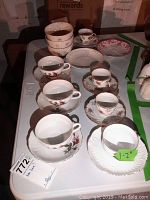 7 assorted teacups with saucers and 3 bowls shown on table. Variety of floral designs and white bowls.