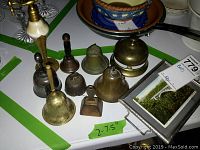 Photo shows 8 bells of various sizes and materials including brass and metal, arranged on a table with a small framed photo visible on the right side.