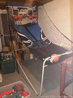 Wide view of the MD Sports 3-in-1 basketball shooter game showing the dual hoops, black mesh side nets, blue and black fabric ramp, and ball return setup in a basement environment.