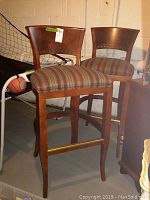 Two wooden bar stools with striped cushions and curved backrests shown at an angle inside basement area with sports equipment nearby.