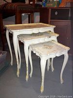 Set of three white nesting tables with floral painted tops, curved legs, and gold detailing shown from side angle