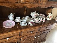 Wide shot of the full tea set collection displayed on a wooden cabinet, showing various plates, cups, teapots, and the pitcher.