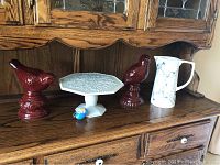 Side view of five items on wooden surface, including two red bird figurines, white decorative plate on pedestal, white marbled pitcher, and small blue blown glass egg ornament.