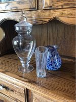 Three glass items displayed on wood surface: a tall lidded clear glass apothecary jar, a blue and white speckled vase, and a clear Lenox crystal vase with scalloped rim.