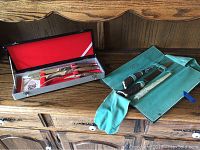Overview of two carving sets displayed on a wooden surface, one in a metal case and the other in a green fabric pouch