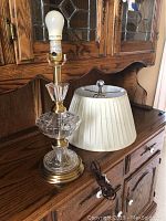 Full view of the crystal table lamp without the shade, showing the clear faceted crystal base with brass hardware and attached light bulb, alongside the lampshade and power cord on wooden furniture.