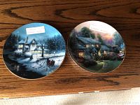 Two Thomas Kinkade collector plates on a wooden surface, showing detailed cottage scenes with winter and garden themes.