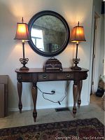 Full view of dark wood console table with drawer, holding the two matching lamps and ceramic chest, with round smoked glass mirror mounted on wall above.