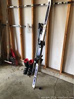Photo showing one ski leaning against a wall with two ski poles beside a pair of boots on a concrete floor