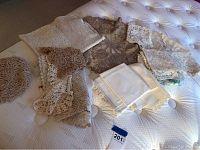 Photo showing an assortment of crocheted and lace linens including pillowcases on a white tufted surface.