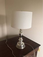 Full view of the silver table lamp on a wooden surface showing the white fabric shade and metallic reflective base with power cord visible.