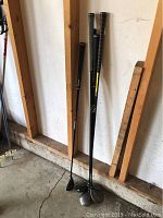 Five golf clubs leaning against a wall in a garage or storage area, showing their grips and club heads.