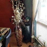 Photo showing tall mosaic tile patterned vase with dried foliage, black ceramic stand holding a glass bowl with decorative spheres beside a window and chair.
