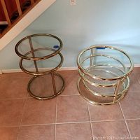 Two three-level glass side tables with gold-colored metal frames placed on tiled floor against wall, showing both tables side by side from above angle.